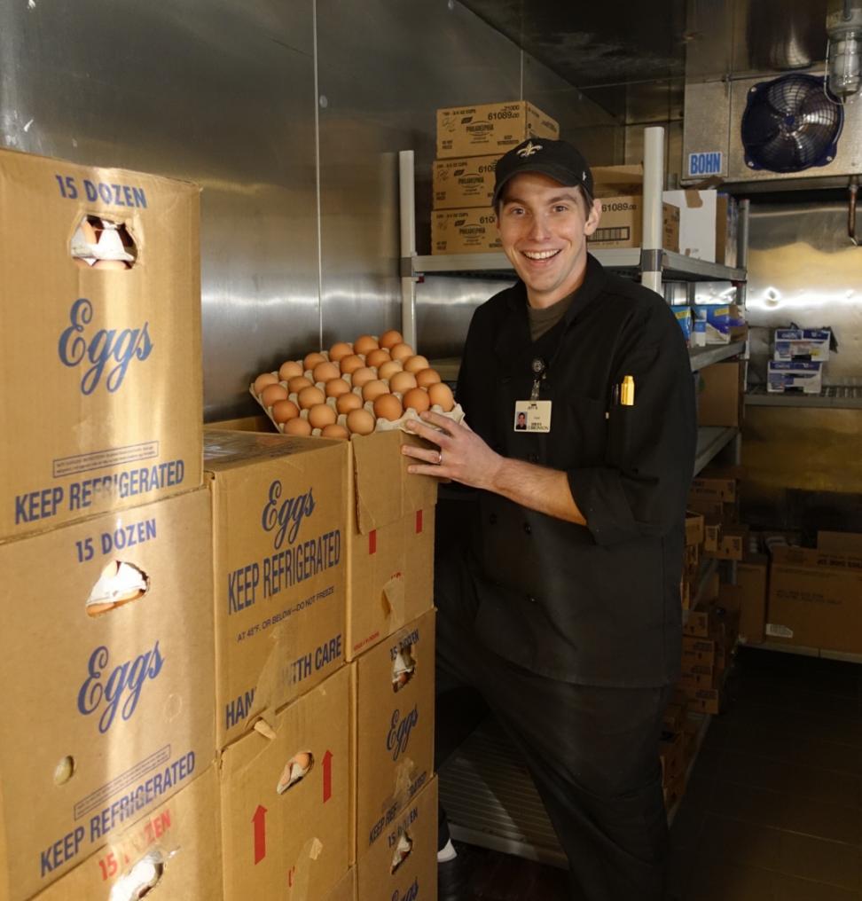 A man in a chef jacket and hat holds 2.5 dozen eggs in an industrial kitchen.